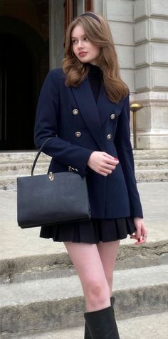 Woman in a navy blazer and black pleated skirt holding a handbag outdoors