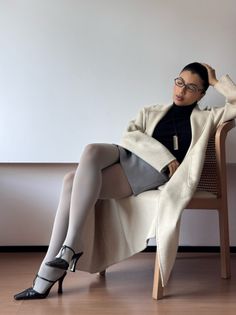 Woman in a long coat, turtleneck, mini skirt, and white tights sitting on a chair