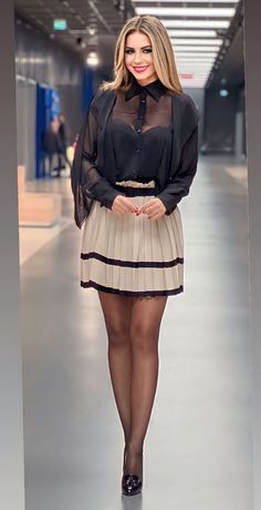 Woman in a sheer black blouse and pleated skirt smiling indoors
