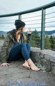Woman in a beanie and jacket sitting barefoot outdoors, making a peace sign