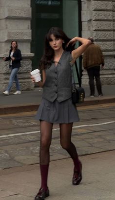 Woman in a gray vest and pleated skirt walking on a street with coffee