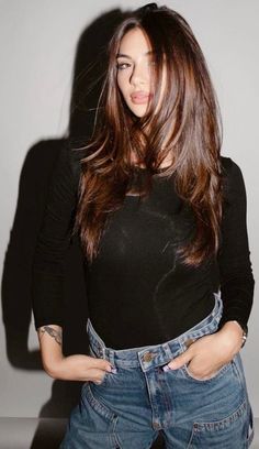 Woman with long brown hair wearing a black shirt and jeans against a gray wall