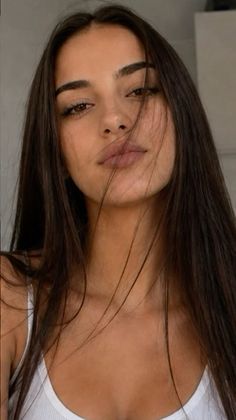 Woman with long dark hair wearing a white tank top, looking directly at the camera
