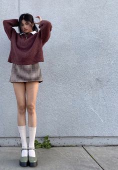 Woman in a cropped sweater and plaid skirt posing against a wall
