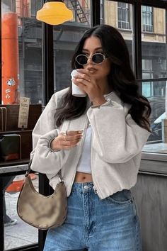 Woman in sunglasses, crop top, and jeans holding a coffee cup indoors