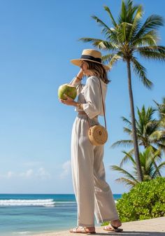 Woman in a straw hat and linen outfit holding a coconut drink on a beach