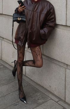Person in a brown leather jacket and lace tights leaning against a wall