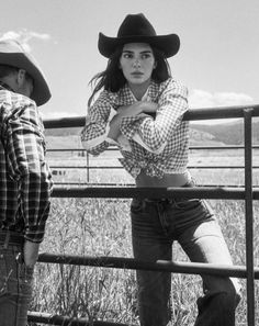Woman in a cowboy hat and tied gingham shirt leaning on a fence in the countryside