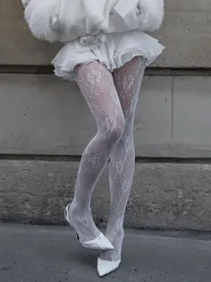 Person wearing white lace tights, a white skirt, and white heels against a concrete wall