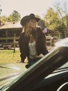 Woman in a cowboy hat and corset leaning on a car in a countryside setting