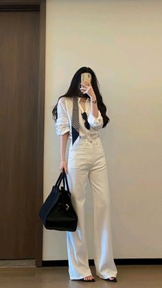 Woman in a white shirt and pants holding a black handbag, taking a mirror selfie