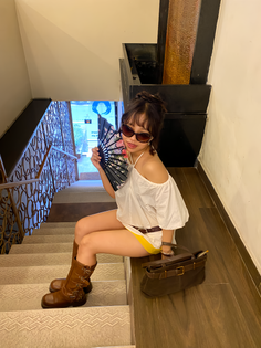 Woman in an off-shoulder top and yellow shorts sitting on stairs with a fan