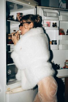 Woman in a white fur coat and lace tights eating from a refrigerator