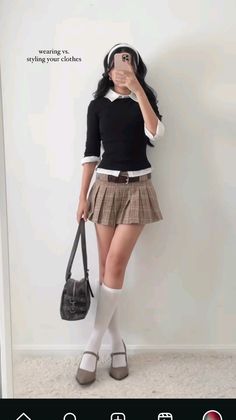 Person in a plaid skirt and black top holding a phone, standing indoors