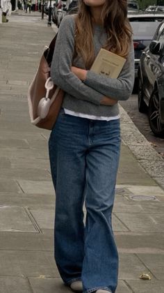 Person in a grey sweater and blue jeans holding a book on a street