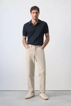 Man in a dark polo shirt and beige pants standing against a plain background