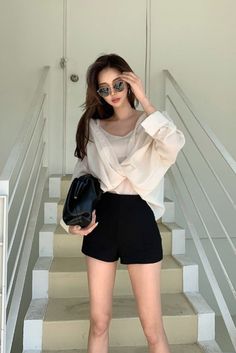 Woman in sunglasses, white shirt, and black shorts holding a handbag on stairs