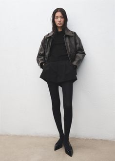Woman in a black leather jacket, shorts, and tights against a white wall