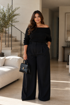Woman in a black off-shoulder top and wide-leg pants holding a handbag indoors
