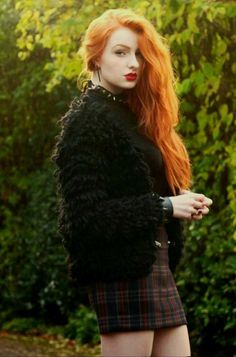 Woman with long red hair and red lipstick wearing a black fuzzy jacket and plaid skirt