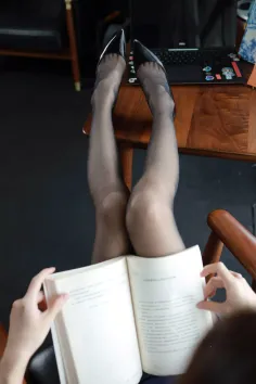 Person in black stockings and high heels with legs up on a table, reading a book