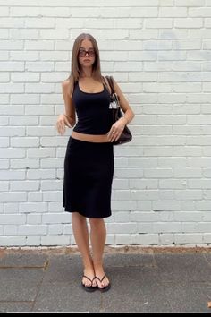 Woman in a black tank top and midi skirt posing against a white brick wall