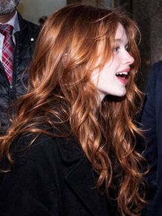 Woman with long wavy red hair in a black coat, smiling in profile