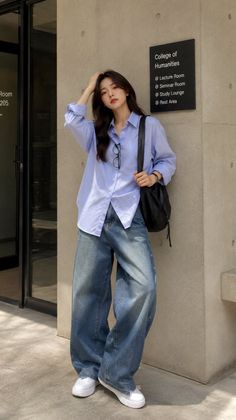 Woman in a blue shirt and wide leg jeans leaning against a wall outdoors