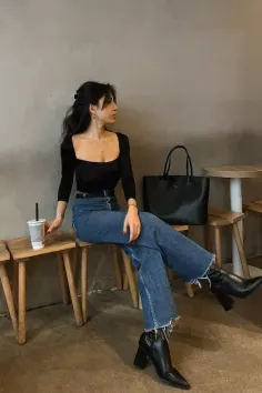 Woman in a black top and blue jeans sitting on a wooden stool indoors