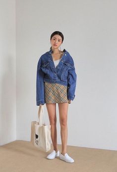 Woman in a denim jacket and plaid skirt holding a tote bag indoors
