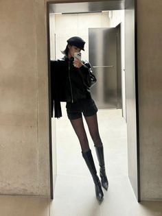 Woman in a black leather jacket, shorts, and knee-high boots taking a mirror selfie
