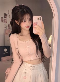 Woman with long dark hair taking a mirror selfie in a pink top and white skirt
