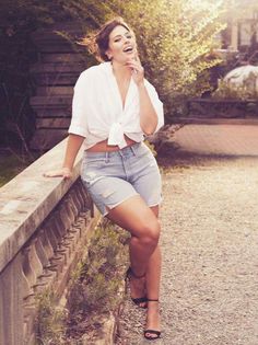 Woman in denim shorts and a white shirt leaning on a stone wall outdoors