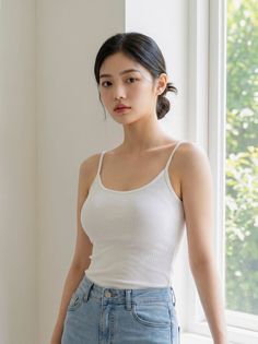 Woman in a white tank top and blue jeans standing by a window