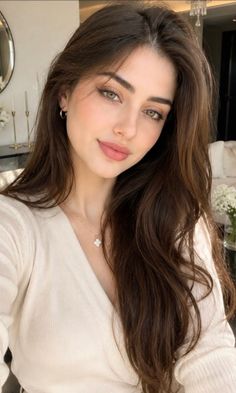 Woman with long brown hair and green eyes wearing a white top and necklace indoors