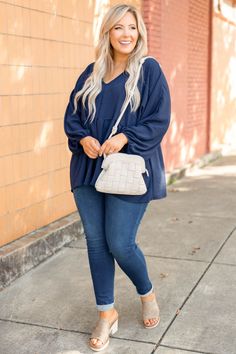 Woman with blonde hair wearing a navy blue top and jeans, holding a crossbody bag outdoors