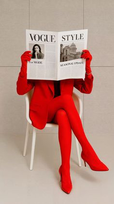 Person in a red suit, gloves, and tights sitting on a chair reading a newspaper
