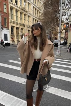 Woman in a beige coat and black shorts standing on a city street crosswalk