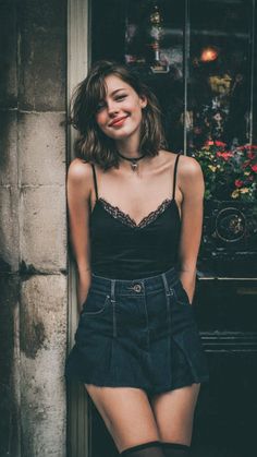 Woman with short hair in a black camisole and denim skirt leaning against a wall