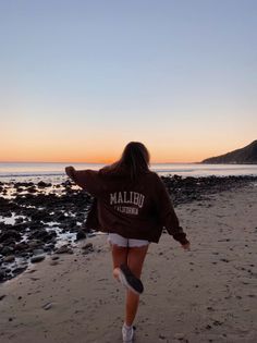 Person in a hoodie and shorts walking on a rocky beach at sunset