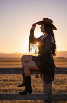 Woman in a fringe dress and cowboy hat sitting on a fence at sunset