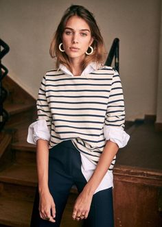 Woman in a striped shirt and dark pants with hoop earrings indoors