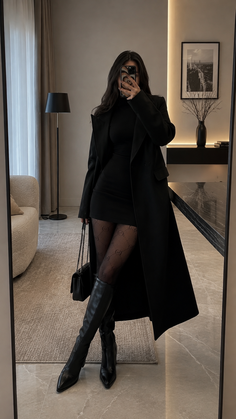 Woman in a black coat and mini dress taking a mirror selfie indoors