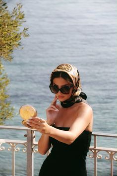 Woman in a silk scarf and sunglasses holding a compact mirror on a balcony overlooking the sea
