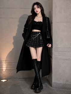 Woman in a black coat, plaid skirt, and knee-high boots standing against a wall