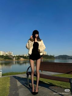 Woman in a fur jacket and black shorts standing by a lake with a cityscape