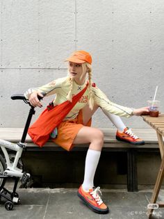 Woman with blonde braids and an orange outfit sitting next to a bicycle
