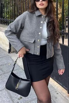 Woman in a gray jacket and black mini skirt holding a black handbag outdoors