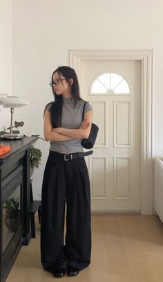 Woman with long hair and glasses wearing a gray top and wide leg pants indoors