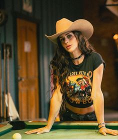Woman in a cowboy hat and crop top leaning on a billiards table